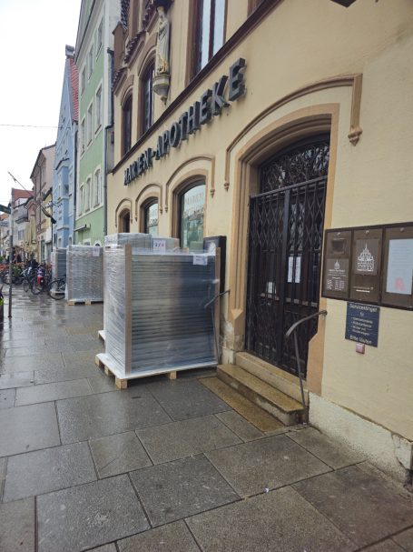 Paletten mit Waren stehen vor einem Gebäudeeingang in einer Stadtstraße.